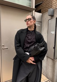 Melnas vilnas jaka pār melnu spiedpogu kreklu, kopā ar plašiem biksēm. Modelis tur melnu ādas zāvju maisiņu.