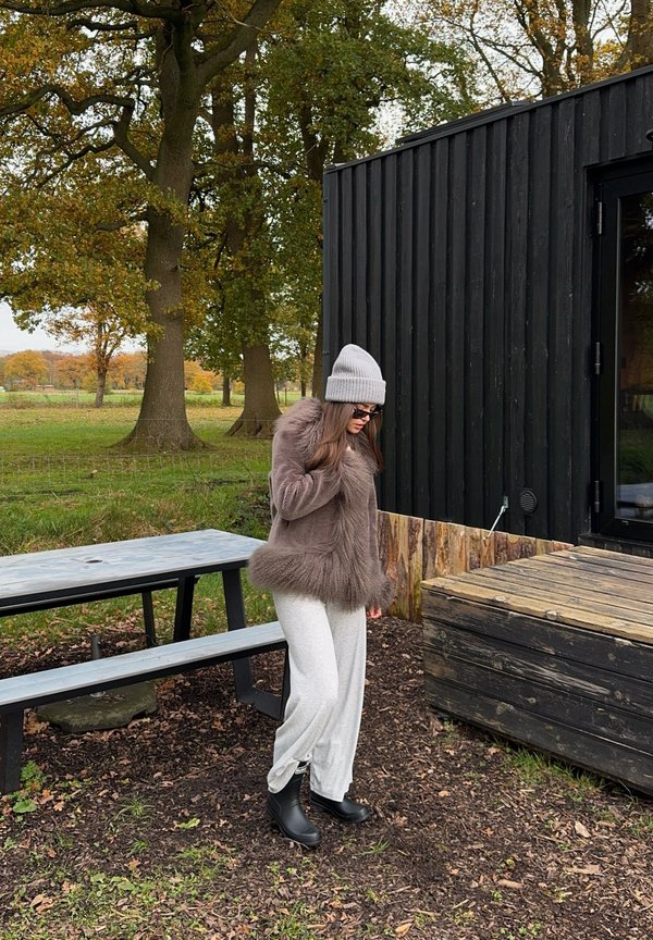 Brown faux fur jacket with a fluffy hem, grey beanie, light grey trousers, and black boots. Outdoors with trees and a wooden structure.