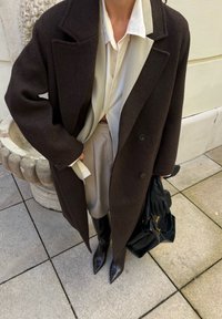 Tmavě hnědý vlněný kabát s velkými klopami, zkombinovaný se smetanovou blůzou a béžovou sukní. Doplněno černými rukavicemi a kabelkou.