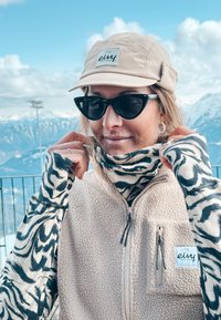 Femme en extérieur dans une région montagneuse portant un gilet en polaire beige, un haut à manches longues à motif animalier, une casquette beige et des lunettes de soleil noires à monture œil de chat.