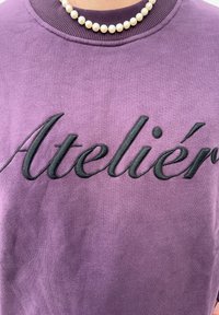 Sweat-shirt violet avec texte noir brodé "Atelier." Encolure côtelée avec texture de tissu lisse, complété par un collier en perles.