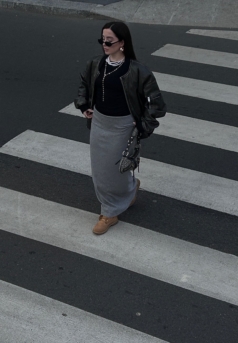 Μαύρο μπουφάν, μαύρη μπλούζα, γκρι μάξι φούστα, καφέ μπότες. Τα αξεσουάρ περιλαμβάνουν κρεμαστά επιχρυσωμένα κολιέ από πέρλες και μικρή τσάντα χειρός. Περπατάει στη διάβαση.