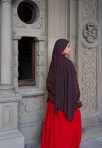 Moteris vilki rudą hidžabą ir ilgą raudoną suknelę, stovėdama šalia puošnios akmeninės architektūros su raižytais raštais ir apvaliu langu.