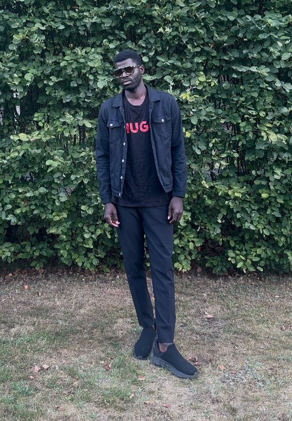 Hombre con gafas de sol, chaqueta negra, pantalones negros y zapatos negros, de pie sobre la hierba frente a arbustos densos y verdes.