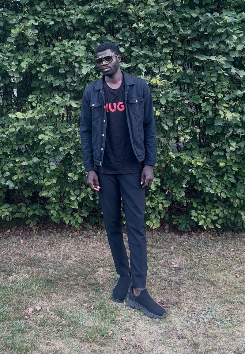 Homme portant des lunettes de soleil, une veste noire, un pantalon noir et des chaussures noires, debout sur l'herbe devant des buissons verts denses.