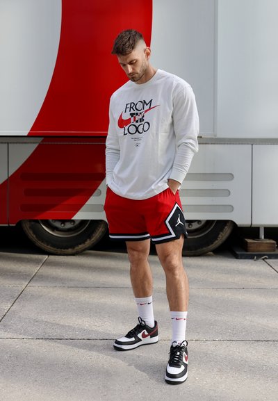 Camisa blanca de manga larga con texto en negro y rojo, pantalones cortos rojos con detalles en negro y blanco, y zapatillas negras con detalles del logo en rojo.