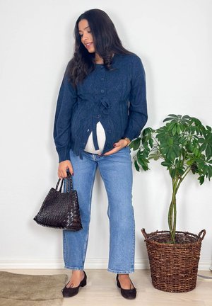 Mujer embarazada con suéter azul y vaqueros sosteniendo un bolso oscuro tejido, de pie en el interior junto a una planta verde en una cesta de mimbre.