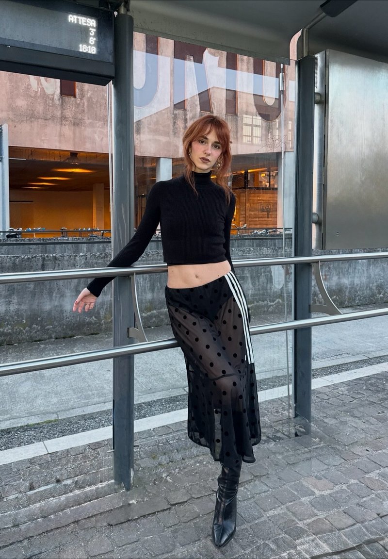 Jeune femme en pull court noir, jupe transparente à pois, et bottes, appuyée contre une rambarde à un arrêt de bus avec un affichage numérique des horaires.