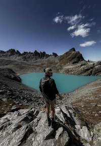 Mägironija, kes kannab mütsi ja seljakotti, seisab kivisel maastikul, vaadates mägijärve, mille vesi on türkiissinine ning taevas on selge ja sinine.