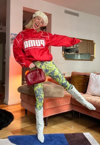 Personne portant un béret beige, une veste rouge Puma, un pantalon jaune à motifs et des bottes blanches, posant joyeusement avec un sac à main rouge dans un salon.
