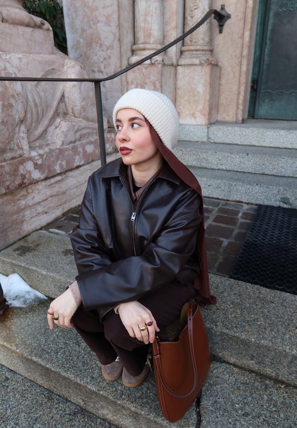 Jeune femme portant un bonnet en maille blanc et une veste en cuir sombre, assise sur des marches en pierre à côté d'un sac à main marron, regardant vers sa gauche.