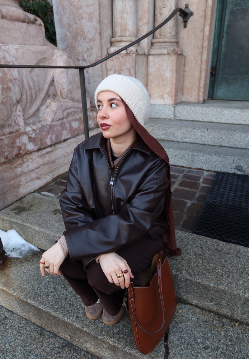 Joven con un gorro de punto blanco y chaqueta de cuero oscura sentada en unos escalones de piedra junto a un bolso marrón, mirando hacia su izquierda.