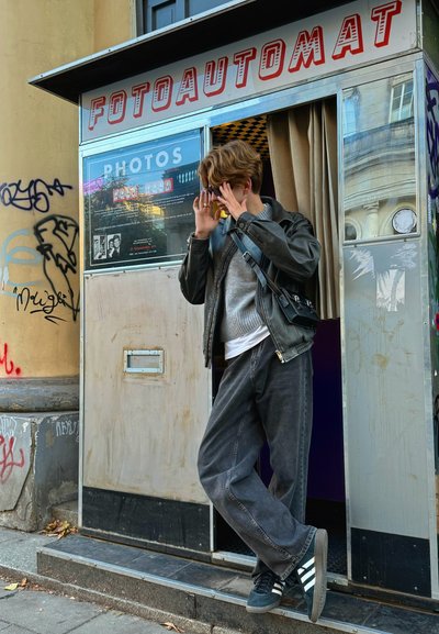 Cabina de fotoautomat con paneles espejados, con un techo de cuadros y letreros. Persona con chaqueta de cuero y ropa gris está de pie en la entrada.
