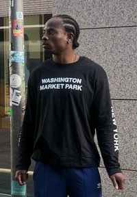 Μαύρο μπλουζάκι με μακρύ μανίκι που γράφει "WASHINGTON MARKET PARK" και "NEW YORK" με λευκά γράμματα. Φοριέται με μπλε αθλητικά παντελόνια.