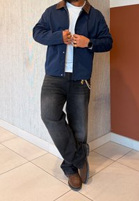 Veste bleu marine avec un col marron, t-shirt blanc, jean foncé et chaussures marron. Comprend des anneaux dorés et un détail de poche à chaîne.