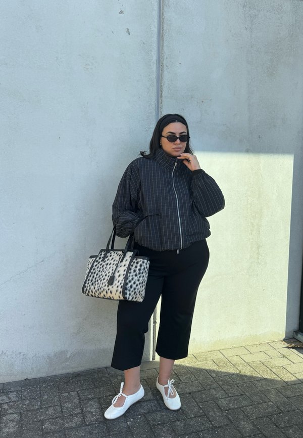 Mujer con chaqueta negra de rayas, pantalones negros, zapatos blancos, gafas de sol, sosteniendo un bolso negro y blanco con manchas frente a una pared gris.
