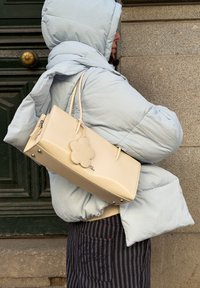 Personne portant une doudoune gris clair à capuche, portant un sac à main rectangulaire beige avec une étiquette en forme de fleur, debout contre un mur en pierre et une porte sombre.