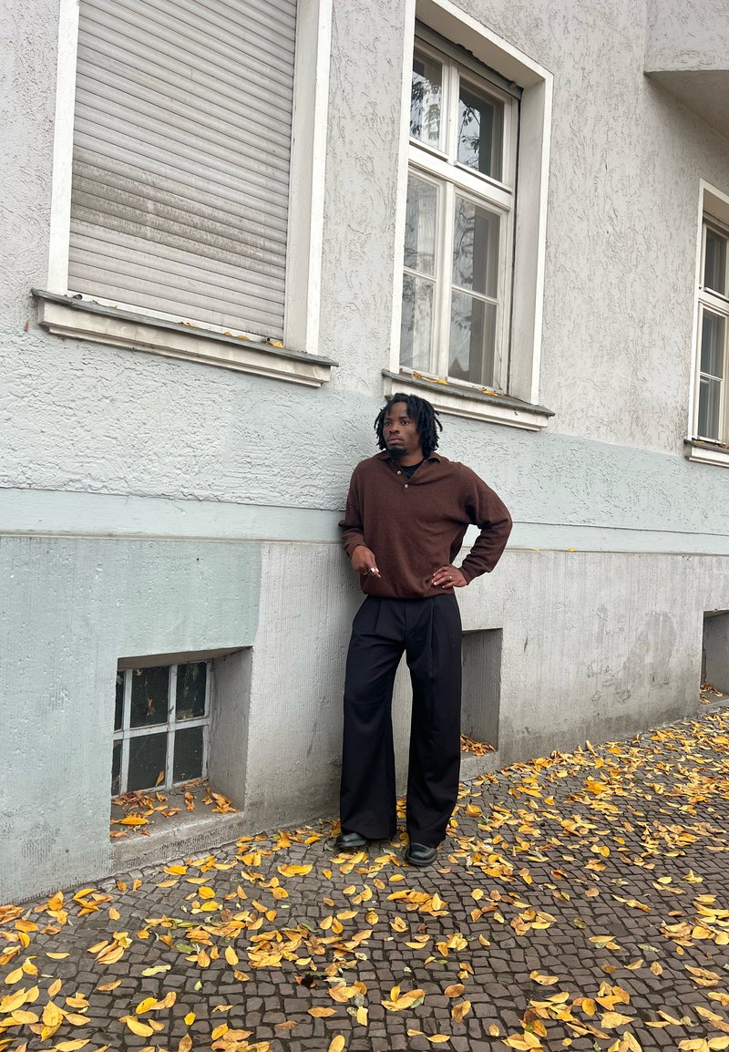 Brauner Strickpullover, schwarze Hose mit weitem Bein, stehend vor einer strukturierten grauen Wand, mit verstreuten gelben Herbstblättern auf dem Boden.