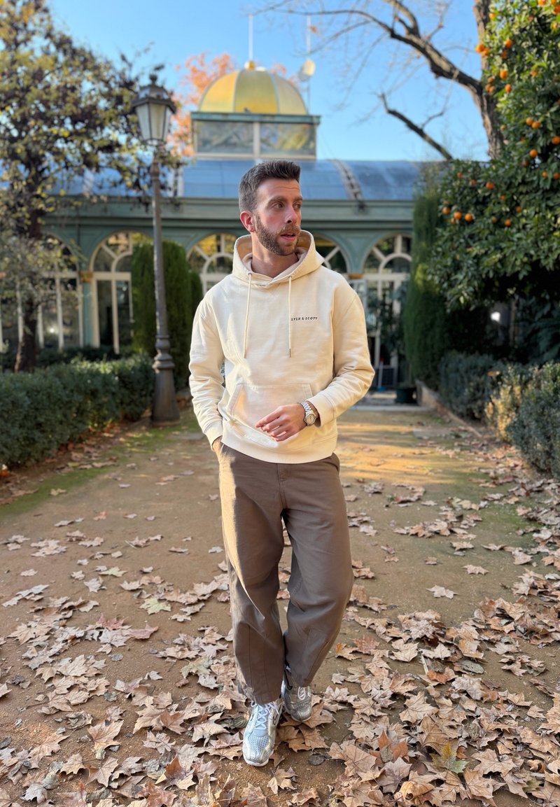 Άνδρας με μπεζ φούτερ με κουκούλα και καφέ παντελόνι περπατά σε μονοπάτι καλυμμένο με φύλλα σε κήπο, με γυάλινο κτήριο και στύλο φωτισμού στο φόντο.