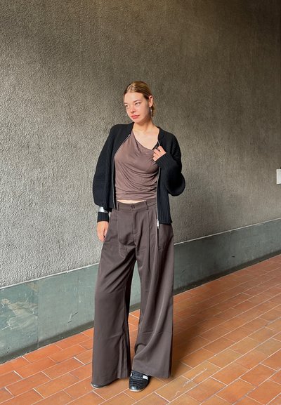 Cárdigan negro de canalé sobre una blusa marrón drapeada, combinado con pantalones de pierna ancha marrones. La modelo lleva zapatos negros sin cordones. Fondo de pared texturizada gris.