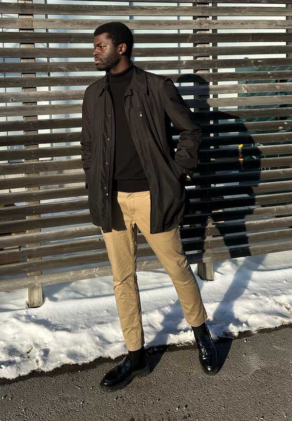 Man in black jacket, black roll-neck, and beige trousers stands on snowy pavement by wooden fence, looking left with hands in pockets.