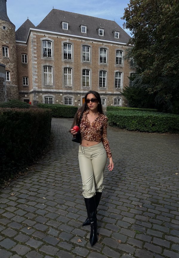 Femme portant des lunettes de soleil et un haut imprimé marche sur un chemin pavé en tenant une pomme rouge devant un bâtiment historique en pierre et de la verdure.