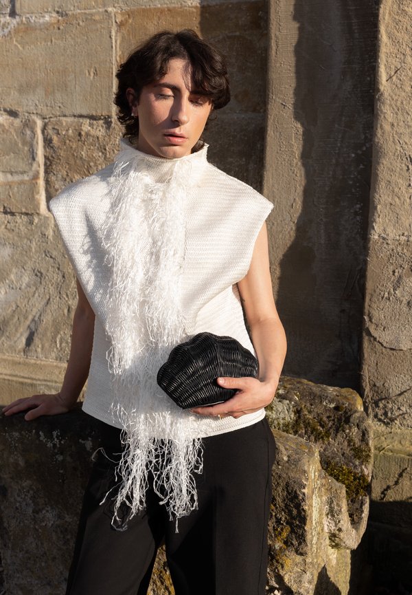 Personne aux cheveux courts et foncés portant un haut blanc sans manches texturé, tenant une pochette noire en forme de coquillage, debout contre un mur en pierre.