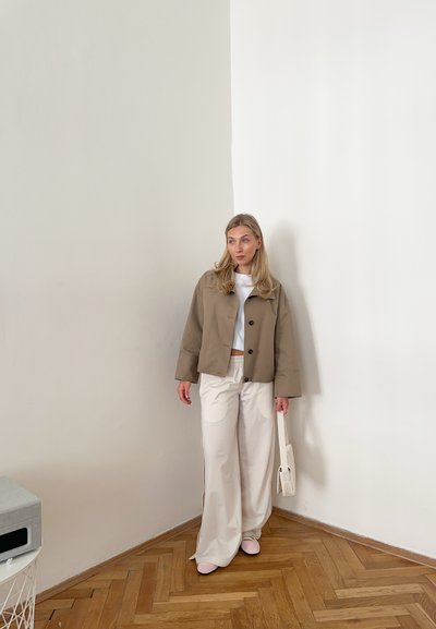 Chaqueta corta beige con botones negros, camiseta blanca, pantalones cremosos de pierna ancha y zapatos rosa claro. De pie en una esquina con suelo de madera.