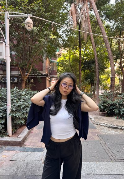 Una mujer con una camisa blanca corta y pantalones negros de pierna ancha está de pie al aire libre, sujetándose el cabello, con vegetación y edificios detrás de ella.
