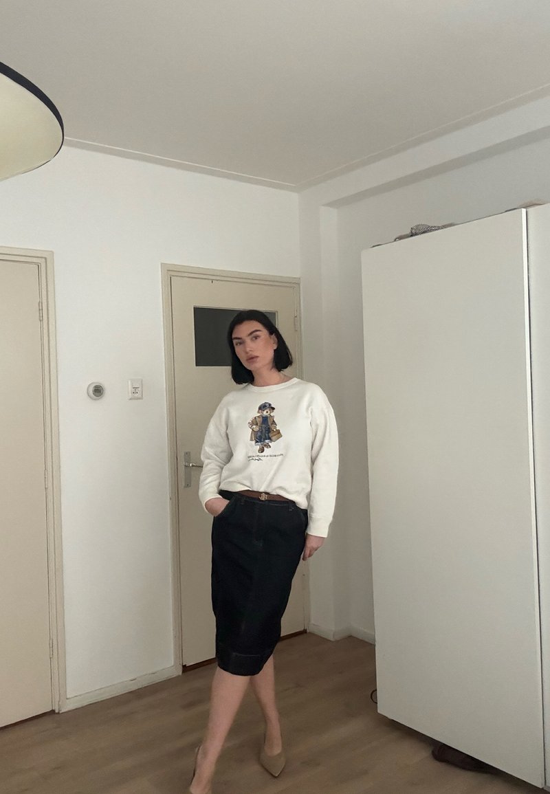 Femme aux cheveux foncés portant un sweat-shirt blanc à motif, une jupe noire jusqu'aux genoux et des talons beiges, debout à l'intérieur près d'une porte fermée et d'une armoire.