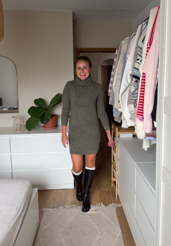 Mujer sonriendo y caminando en el dormitorio vistiendo un vestido de suéter gris y botas negras hasta la rodilla con ribete blanco.