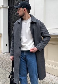 Cappotto di lana grigio scuro con ampio colletto, con bottoni monopetto, indossato sopra una camicia bianca e jeans blu. Zaino nero tenuto sotto il braccio.