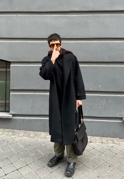 Hombre con abrigo largo negro, bufanda marrón, gafas de sol naranjas, pantalones verdes y botas negras, de pie en la acera de la ciudad ajustándose las gafas de sol con una mano.