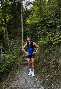 Løper med en blå hydratiseringsvest og svarte shorts, stående på en grussti omgitt av grønt og trær.
