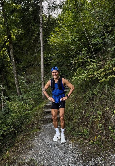Corredor con un chaleco de hidratación azul y pantalones cortos negros, de pie sobre un sendero de grava rodeado de vegetación y árboles.