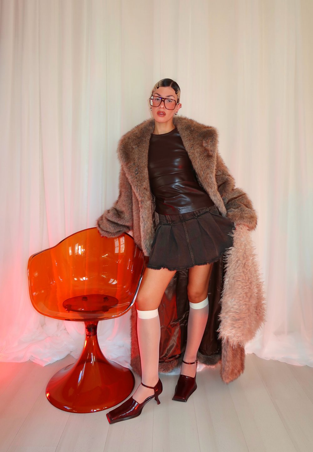 Manteau en fausse fourrure marron, haut en cuir marron, jupe en jean noire, chaussettes blanches montantes jusqu'aux genoux, talons bordeaux, et un fauteuil orange accent.