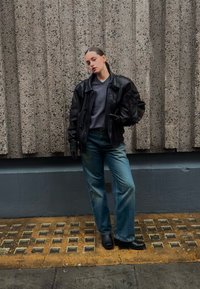 Mustast nahast üle suuruses jakk, hall V-kaelusega kampsun, helesinised laia säärega teksad ja mustad kontsaga saapad tekstuurse betoonseina taustal.