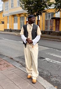 Svart läderväst över en beige rutig skjorta, vita långärmar, cremefärgade byxor och gula träskor; stående mot en gul byggnad.