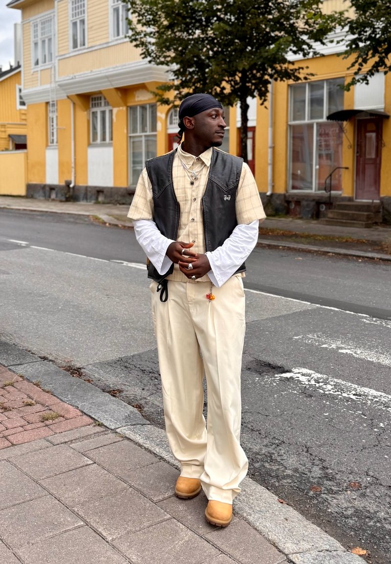 Gilet en cuir noir porté sur une chemise à carreaux beige, manches longues blanches, pantalon crème et sabots jaunes ; debout devant un bâtiment jaune.