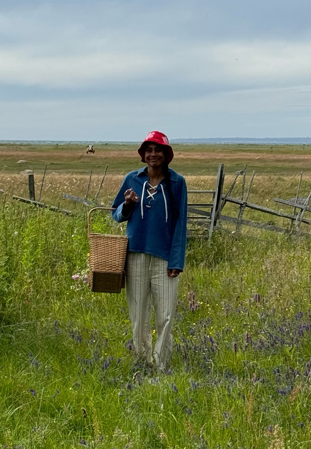 Niebieski sweter z sznurowanym dekoltem, białe spodnie w paski, czerwony kapelusz typu bucket oraz pleciony koszyk w ręku, stojąc na trawiastym polu.