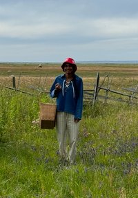 Niebieski sweter z sznurowanym dekoltem, białe spodnie w paski, czerwony kapelusz typu bucket oraz pleciony koszyk w ręku, stojąc na trawiastym polu.