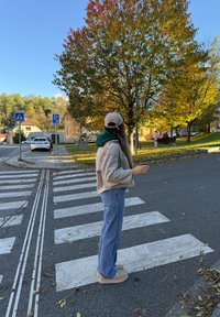 Mulher vestindo roupas casuais de outono, parada numa passagem pedonal, segurando uma chávena, com árvores e carros estacionados ao fundo.