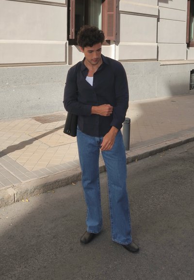 Camisa negra de botones, camiseta blanca debajo, vaqueros de campana azules, zapatos negros, llevando una mochila negra, de pie en una acera.