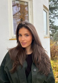 Giovane donna con capelli castani lunghi e giacca verde in piedi all'aperto vicino a un edificio bianco con due finestre e fogliame autunnale.