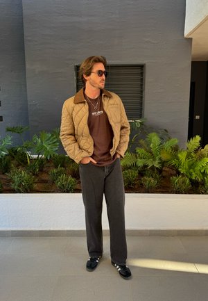 Veste beige matelassée avec col brun, t-shirt graphique brun, jeans foncés amples et baskets noires ; se tenant devant un arrière-plan de feuillage vert.