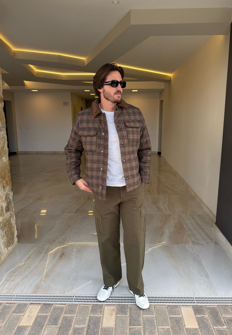 Veste à carreaux marron avec un col beige, t-shirt blanc, pantalon cargo olive et baskets blanches avec des accents verts. Cadre intérieur moderne.