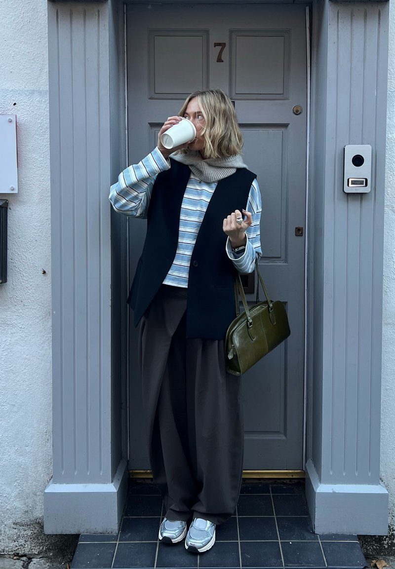 Colete preto sem mangas sobre uma camisa de manga comprida às riscas, calças largas cinzentas, ténis prateados e mala verde. Em pé junto a uma porta.