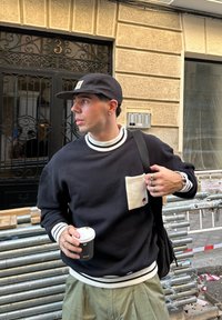 Joven con una gorra negra y un suéter sosteniendo una taza de café y ajustando la correa de la mochila, de pie junto a tuberías de metal afuera de un edificio con el número 3.
