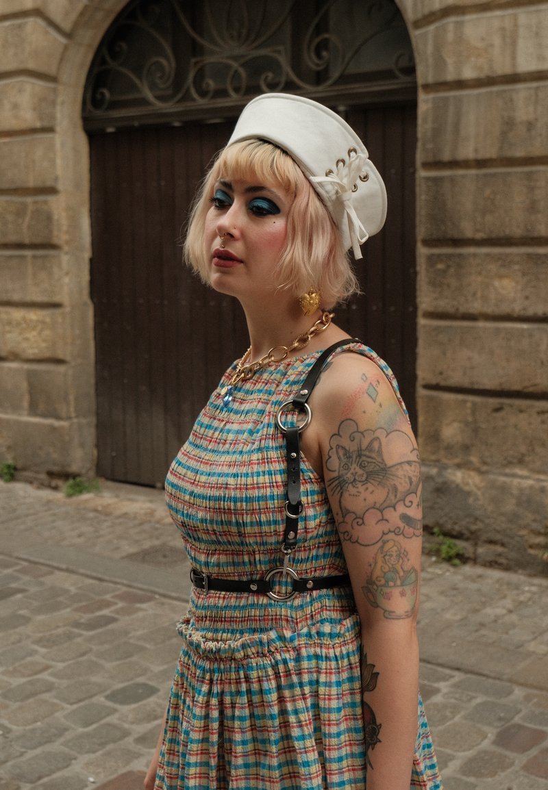 Personne aux cheveux blonds, fard à paupières bleu, chapeau blanc, robe à carreaux, bras tatoué et collier en or se tient dans une rue pavée près de bâtiments en pierre.
