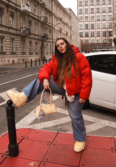 Chaqueta acolchada roja, vaqueros de color claro y zapatos de plataforma amarillos. Bolso beige con un llavero de peluche. Ambiente urbano con edificios.